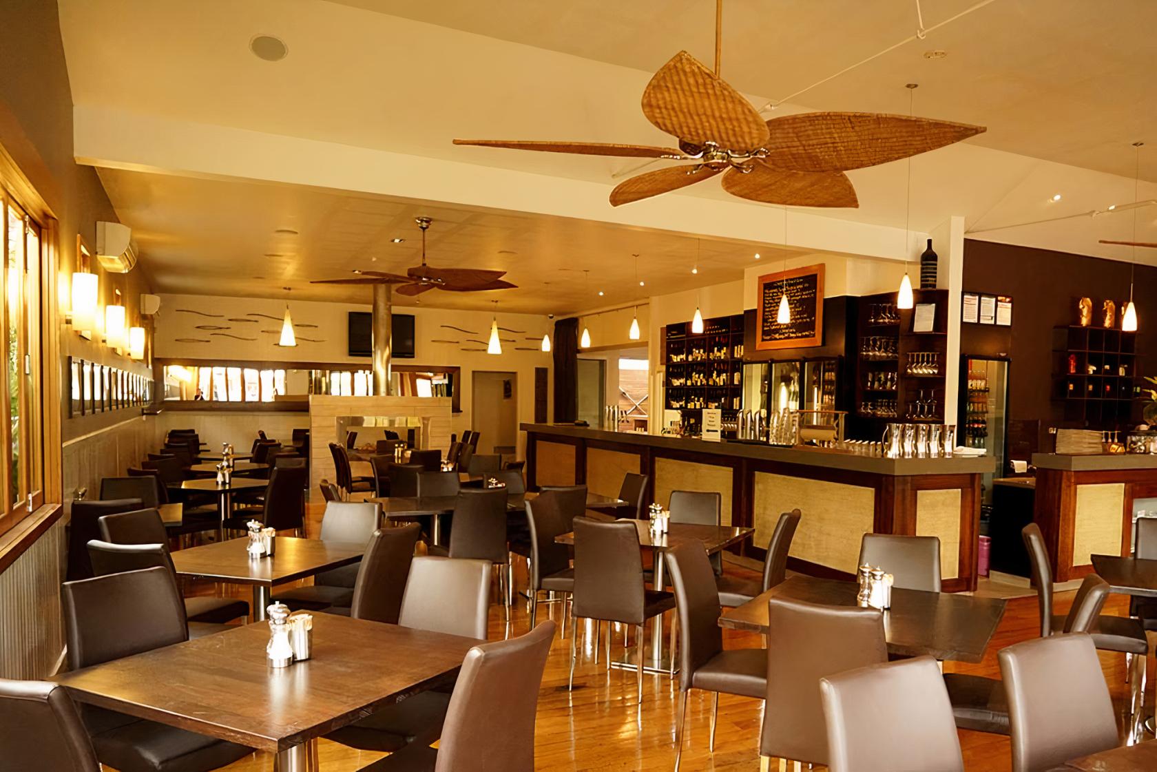 A restaurant with wooden floors, brown leather chairs, pendant lighting, and a central bar at BeachHouse Barwon Heads.