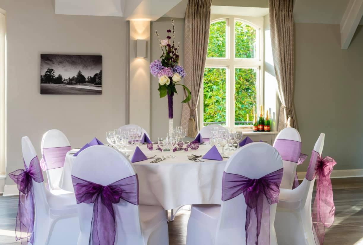 A sports club with round tables dressed in white linens and purple sashes by arched windows at Badgemore Park.