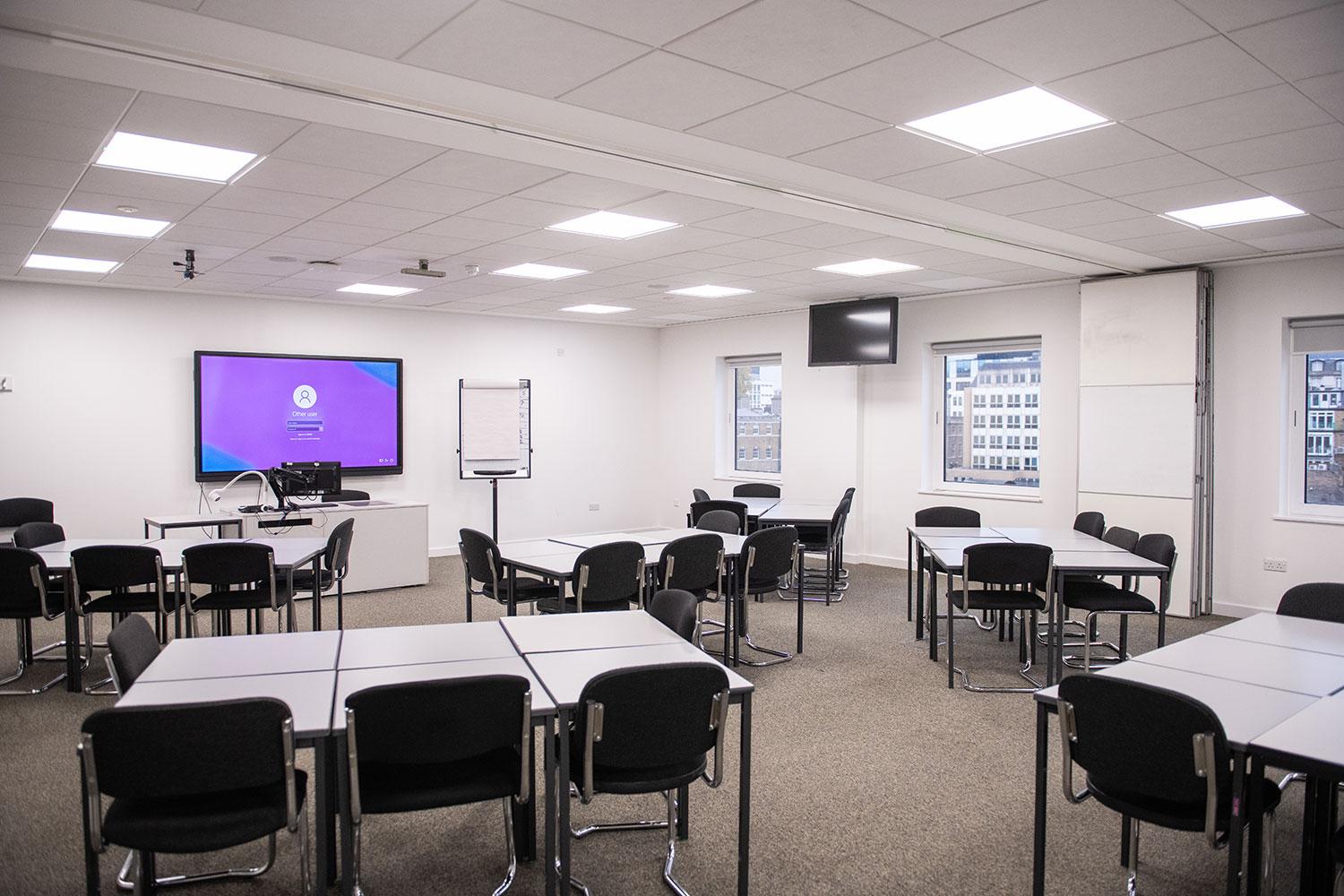 A meeting room with modular tables, black chairs, wall-mounted screens, and large windows at University of Law - London Moorgate.