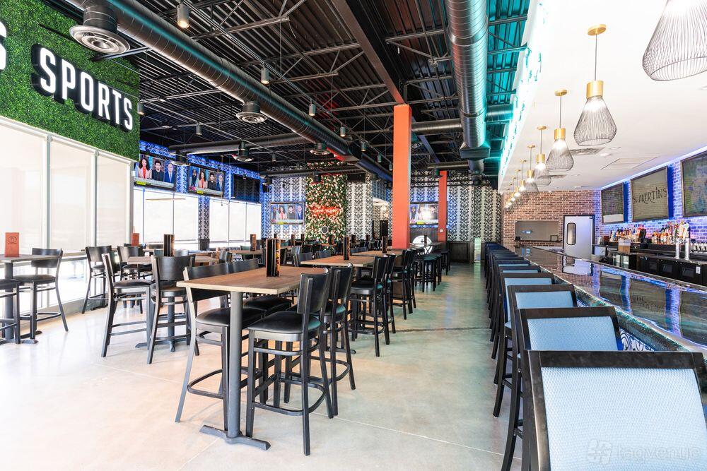 A bar with high-top tables, a long bar counter, industrial ceiling, and large windows at Shakertins- Arlington.