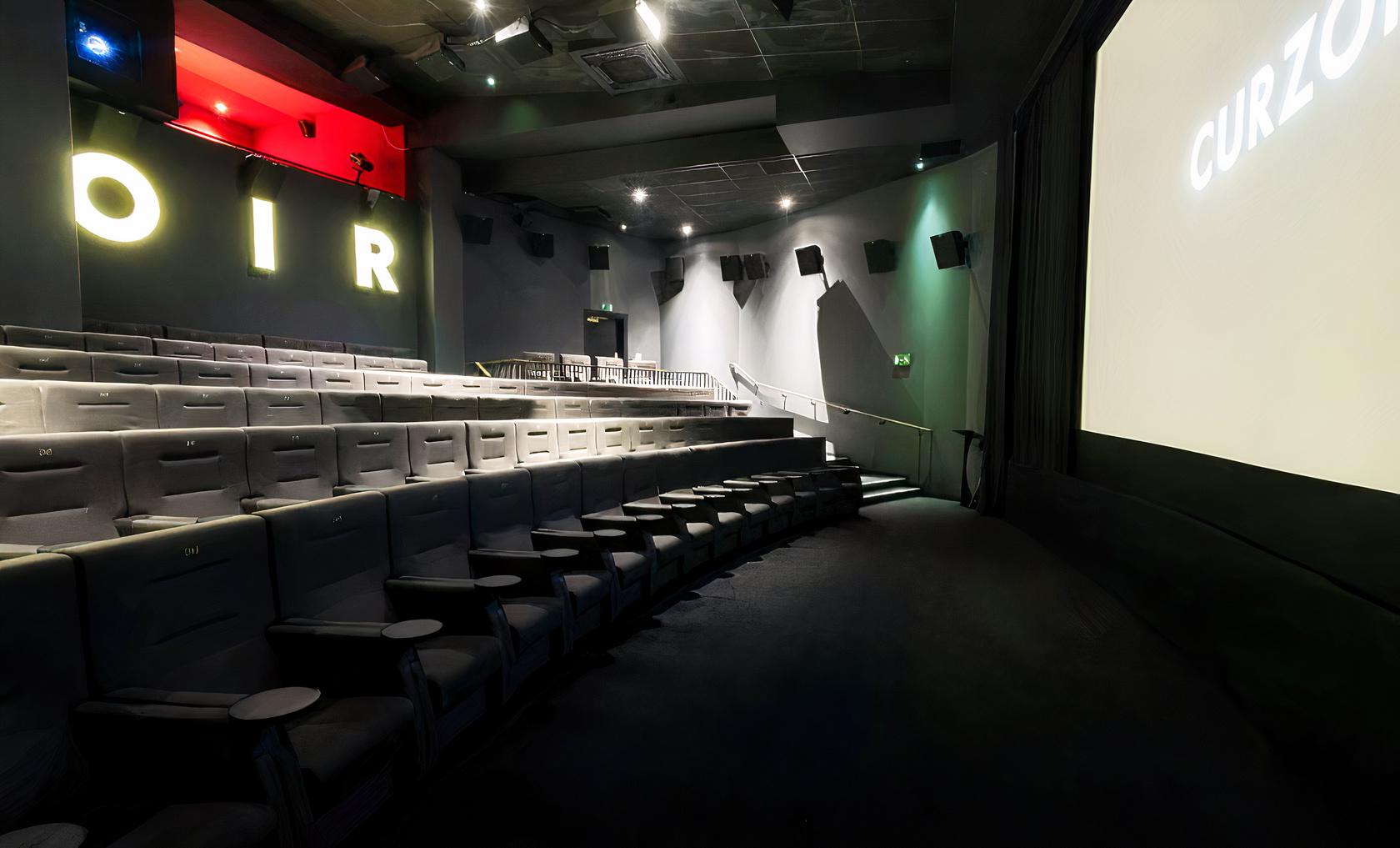 A cinema screening room with tiered grey seats, wall-mounted speakers, and illuminated signage at Curzon Bloomsbury.