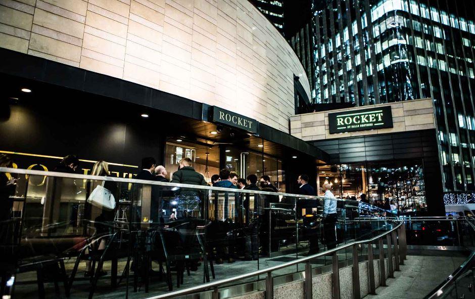 A restaurant with outdoor seating on a glass-railed patio and illuminated signage at Rocket.
