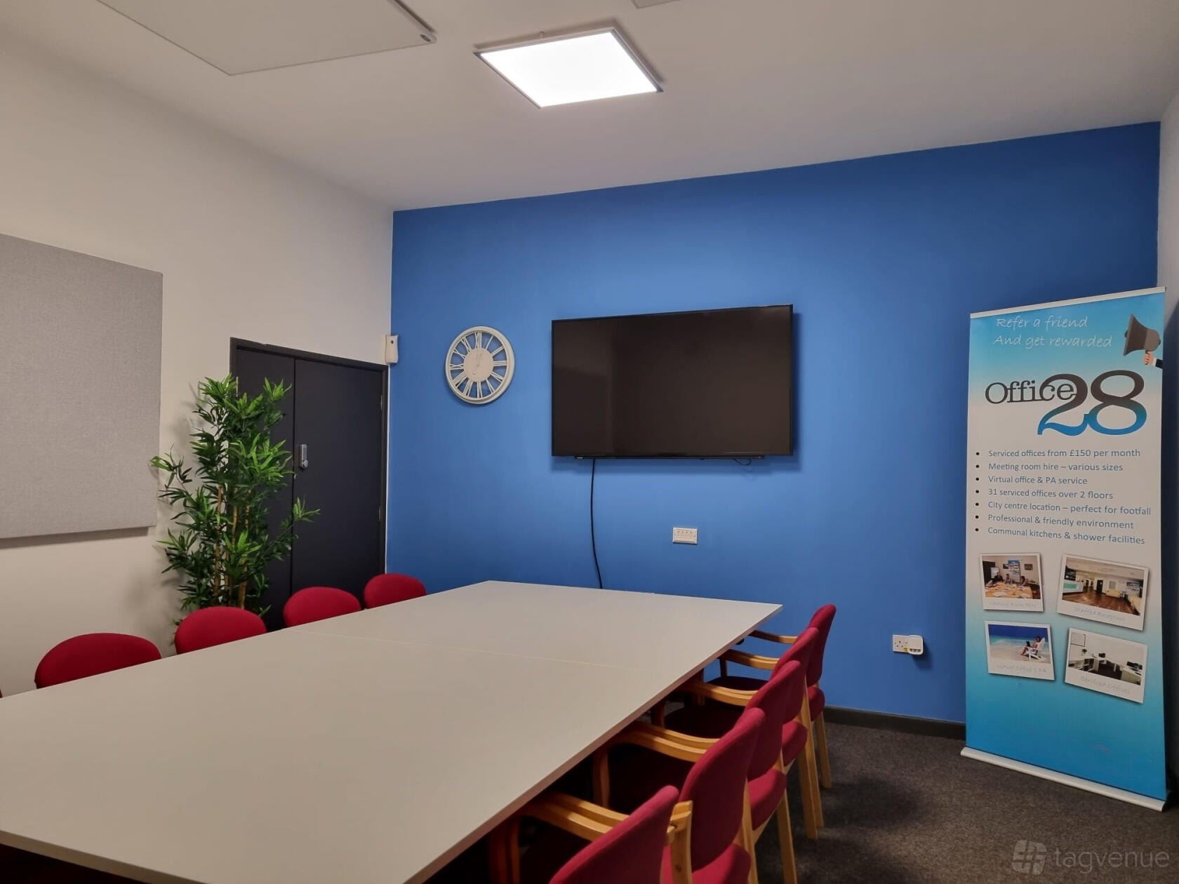 A meeting room with a rectangular table, red chairs, blue accent wall, wall-mounted TV, and indoor plant at Office 28 - Serviced Offices.
