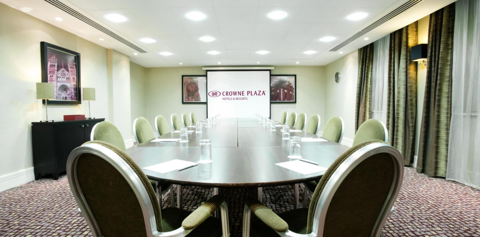 A hotel meeting room with a large oval conference table, green chairs, and a projector screen at Crowne Plaza London Ealing.