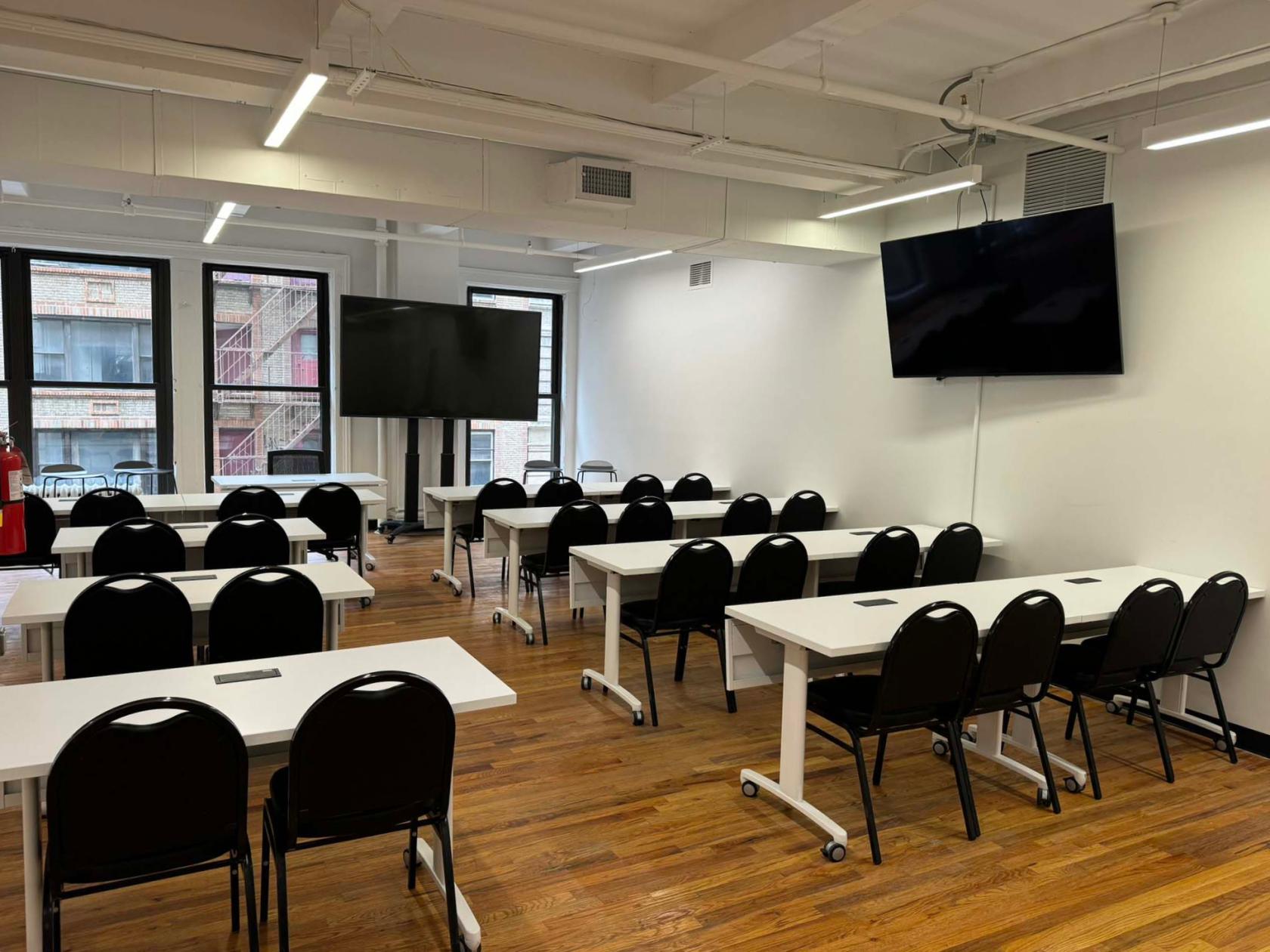 A training room with rows of black chairs, white tables, large screens, and windows at NYC Seminar & Conference Center.