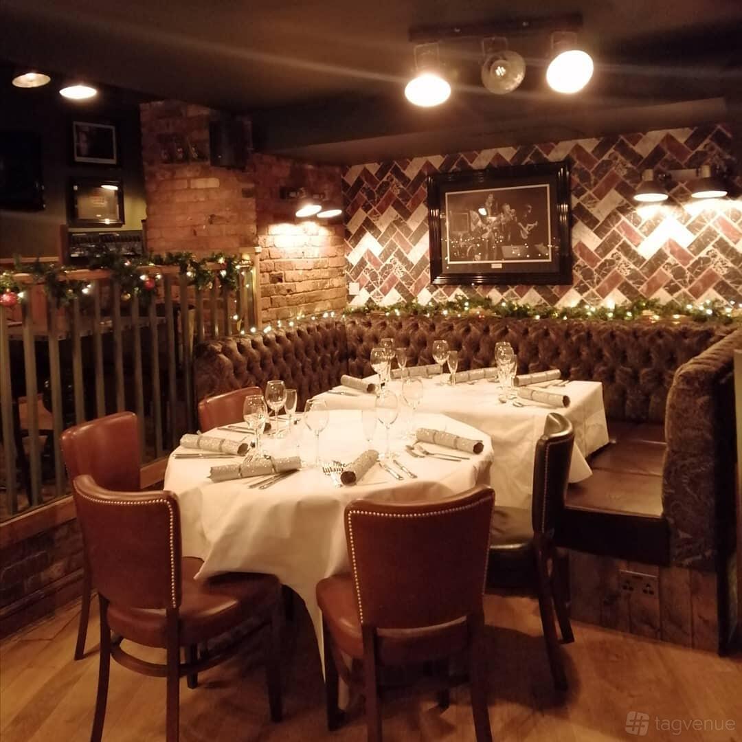 A basement pub area with tufted leather banquette seating, exposed brick walls, and round tables with white linens at The Spice of Life - Soho.