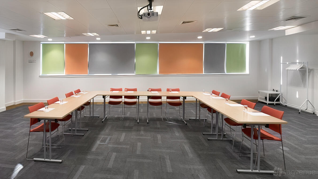 A conference centre room with U-shaped tables, orange chairs, and colorful window blinds at The Studio Glasgow.