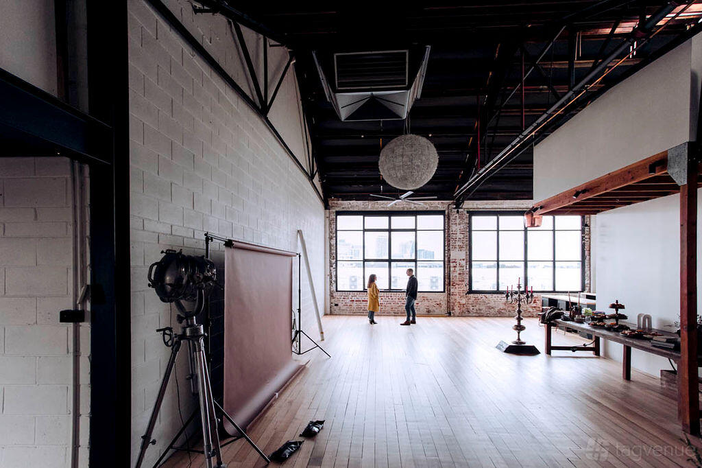 A photo studio with large industrial windows, exposed brick walls, wood floors, and lighting equipment at La Fabrica NY Loft video & Photo Studio.
