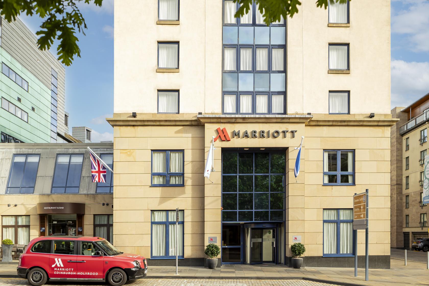 A hotel entrance with large windows, flagpoles, and a red branded taxi at Edinburgh Marriott Hotel Holyrood.