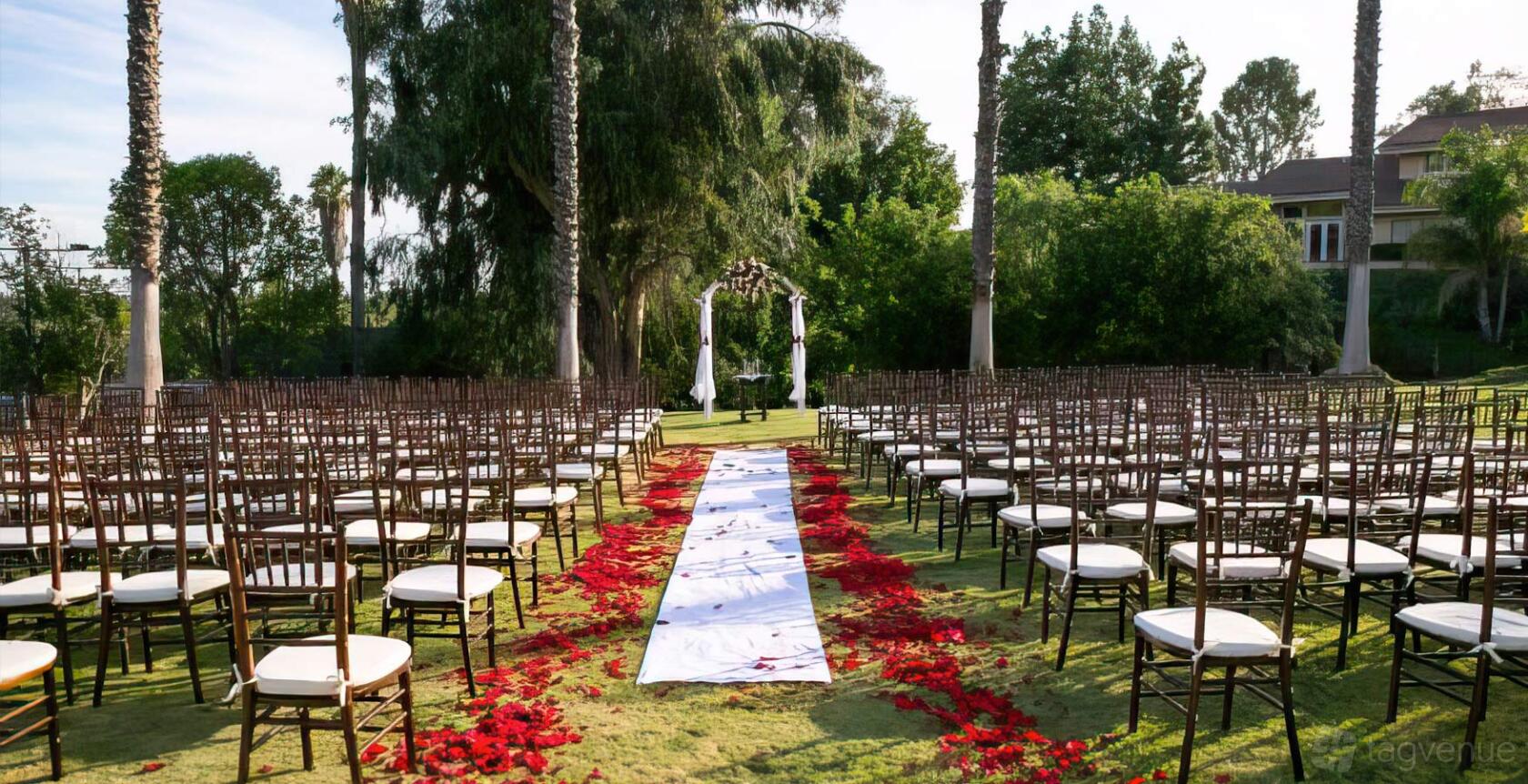 An outdoor event venue with rows of chairs, a rose petal-lined aisle, and a ceremonial arch at Tustin Hills Racquet Club - Legacy Ballroom.