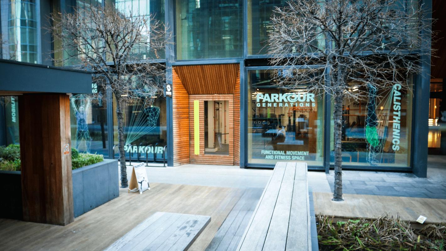A sports hall exterior with large floor-to-ceiling windows, leafless trees, and wood-accented entry at Parkour Generations London.