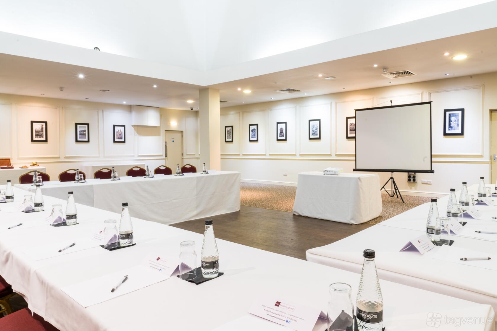 An event space in a hotel with white conference tables arranged in a U-shape, projection screen, and framed wall art at Mosborough Hall Hotel.