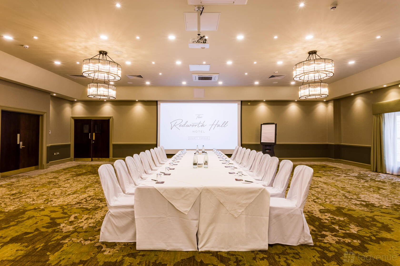 A function room with a large U-shaped conference table, white chair covers, chandeliers, and a projector screen at Redworth Hall Hotel.