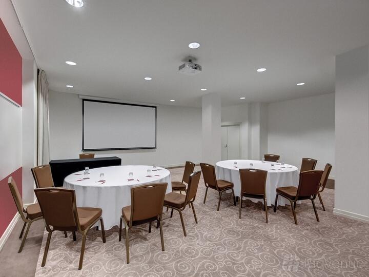A hotel meeting room with round tables covered in white linens, brown chairs, and a ceiling-mounted projector at Rendezvous Hotel Melbourne.