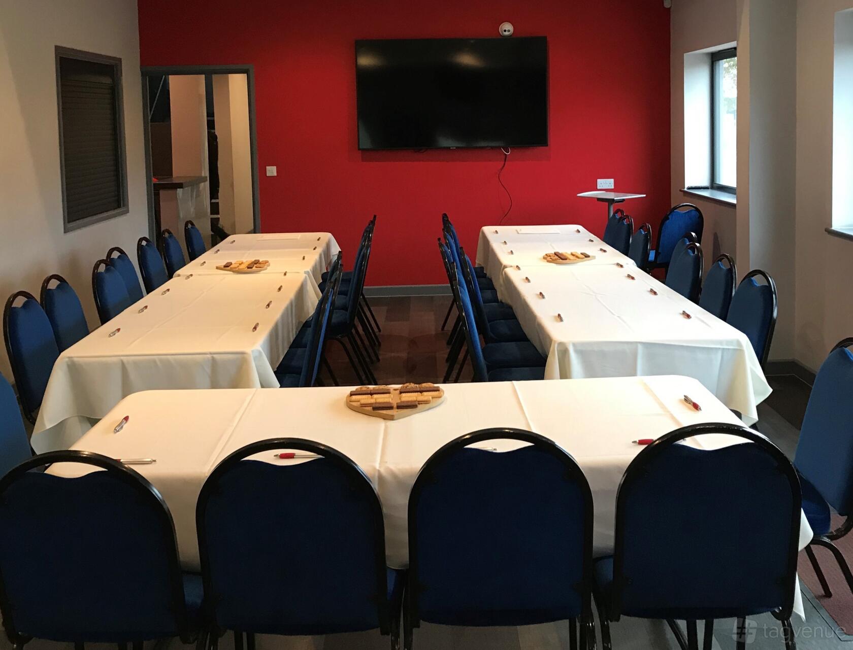 A stadium meeting room with U-shaped tables covered in white linens and blue chairs at Whilton Mill.