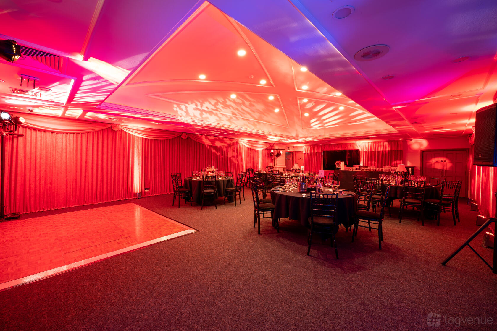 A function room with colored uplighting, round tables with dark chairs, and a wooden dance floor at The Brewery.