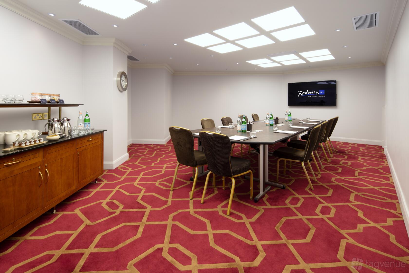 A meeting room with a long conference table, dark chairs, red geometric carpet, and wall-mounted TV at Radisson Blu Hotel, London Heathrow.