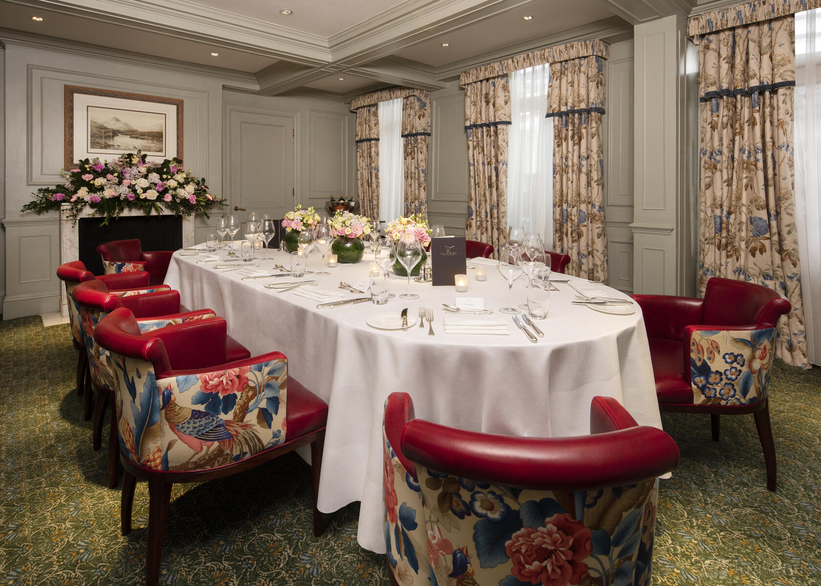 A hotel dining room with an oval table draped in white linens, red upholstered chairs, and floral curtains at The Stafford London.