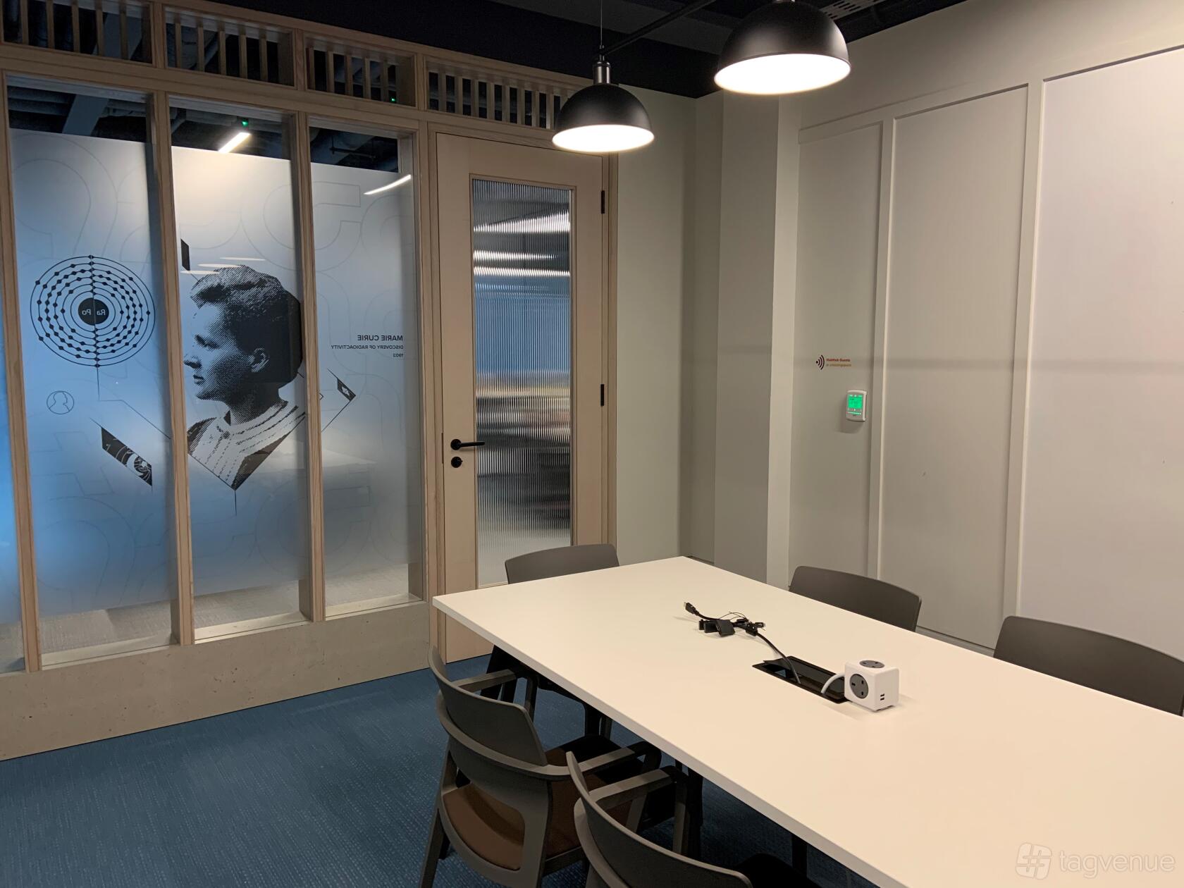 A boardroom with a white table, modern chairs, and a glass wall featuring Marie Curie graphics at HubHub 20 Farringdon Street.