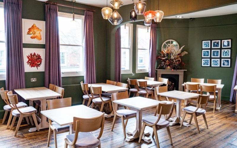 A pub dining room with wooden floors, light wood tables and chairs, pendant lighting, and large windows at The Crossing Pub.