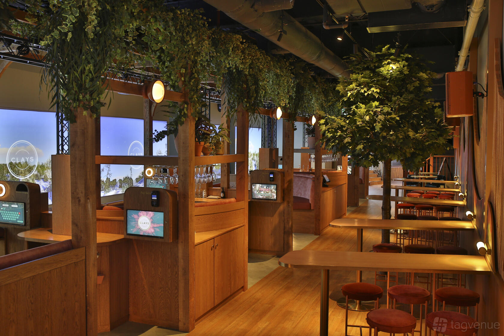A bar interior with wooden booths, digital gaming screens, hanging greenery, and high tables at Clays Canary Wharf.