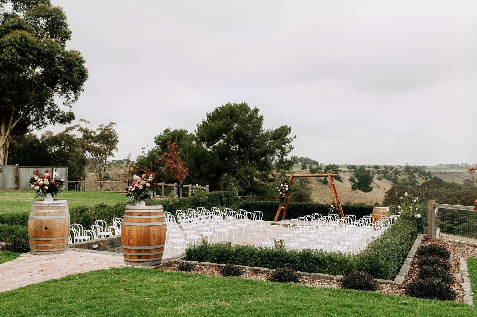 A barn venue with rows of white chairs, floral arrangements on wine barrels, and a wooden arbor at Warrawong Estate.