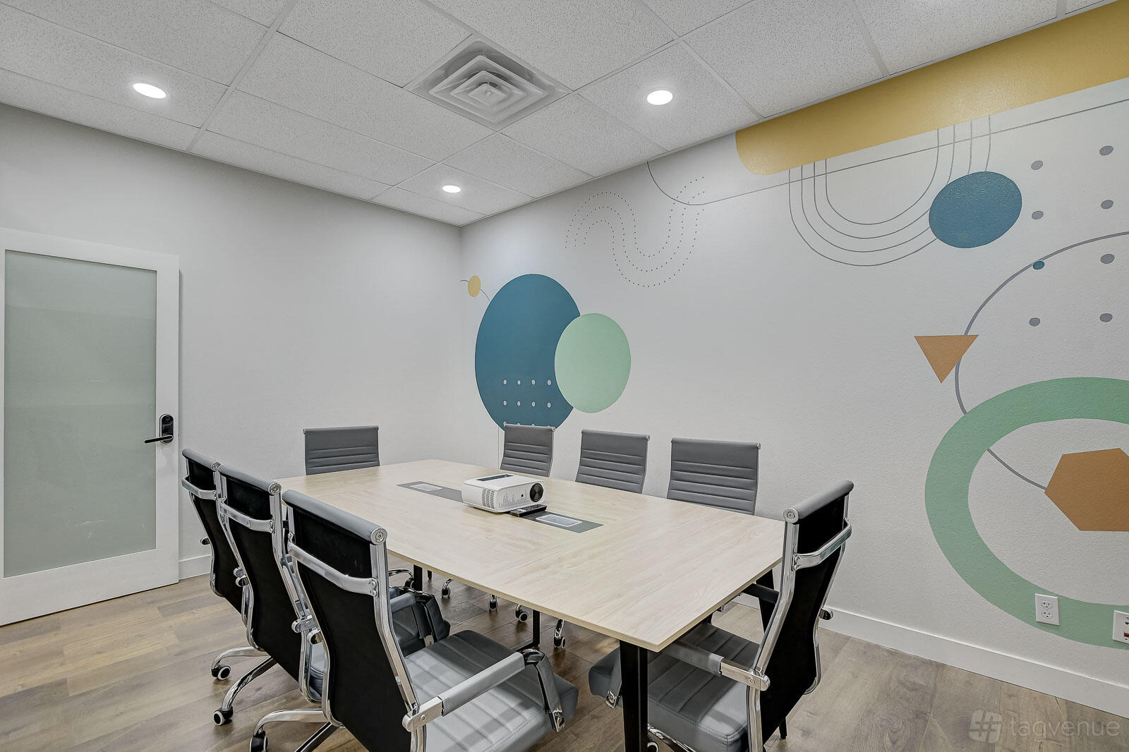 A meeting room with a long wooden table, gray swivel chairs, and geometric wall art at Incubase Workspace.