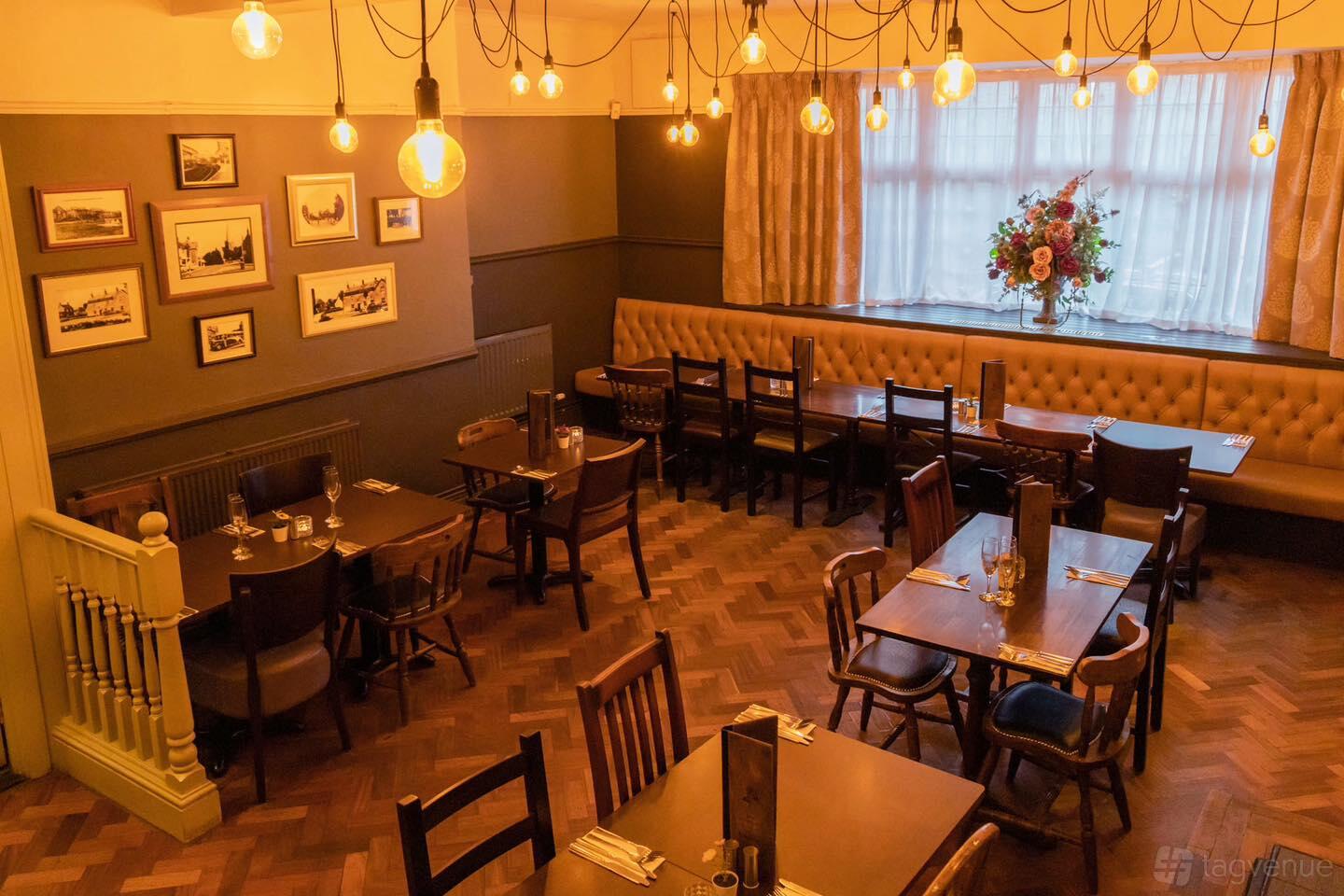 An event space with wooden tables, hanging Edison bulbs, and a banquette under a large window at The Grapevine Brasserie.