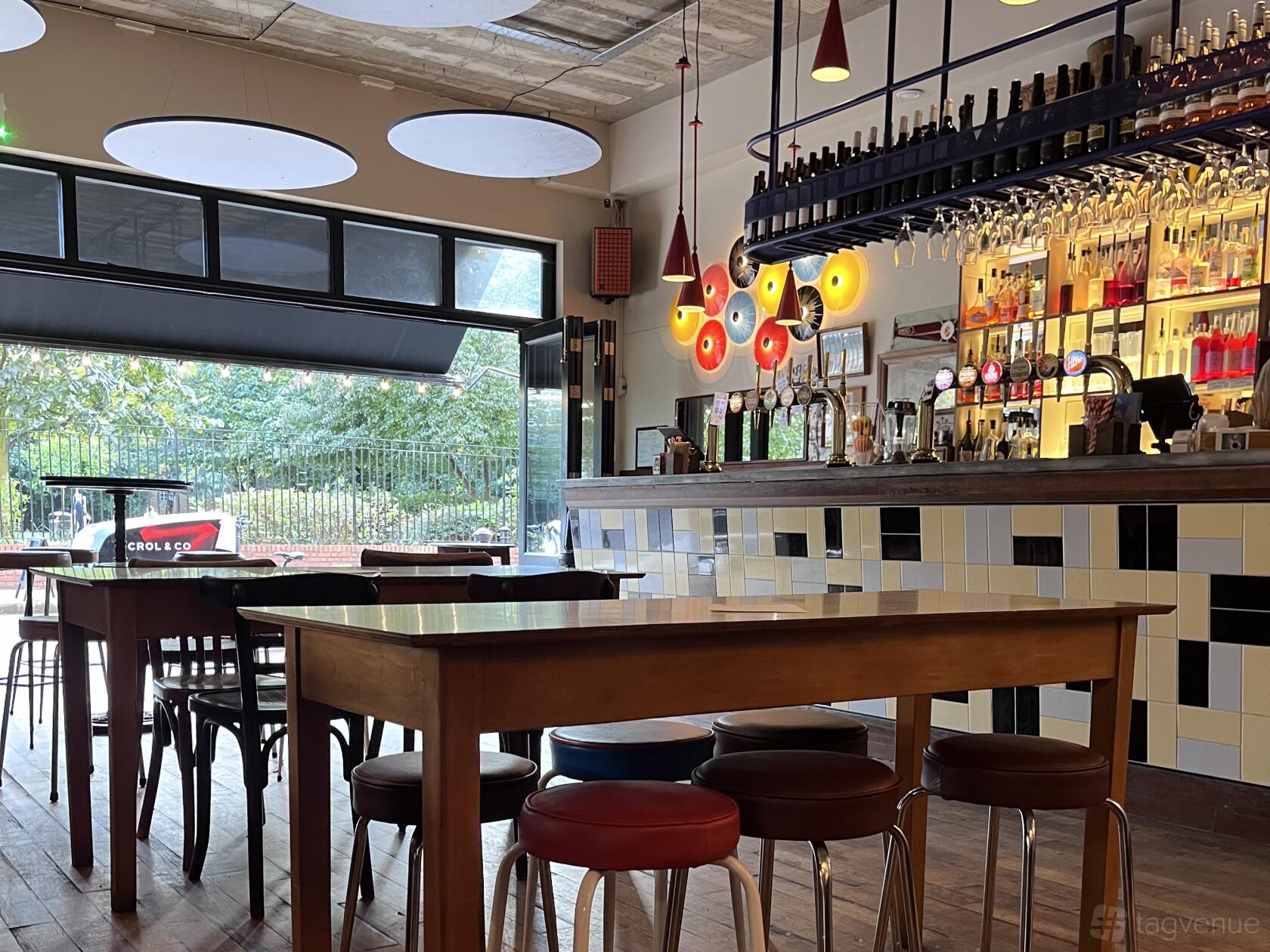A bar with a tiled counter, colorful pendant lights, and wooden tables near large front windows at Crol & Co Bermondsey Street.