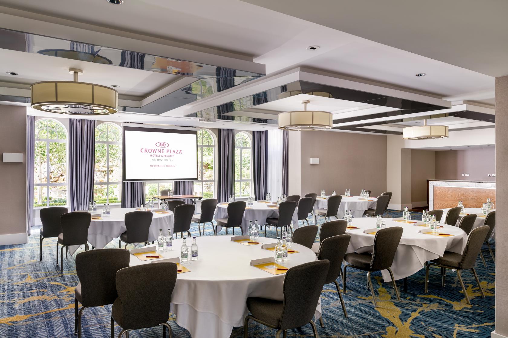 An event space with round tables, bottled water, large windows, and a projector screen at Crowne Plaza Gerrards Cross.