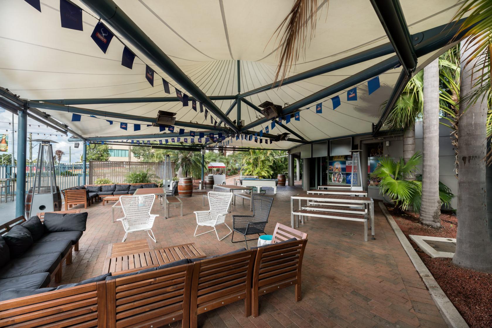 An outdoor hotel space with a large canopy roof, brick flooring, wooden benches, and palm trees at Rydges Parramatta.