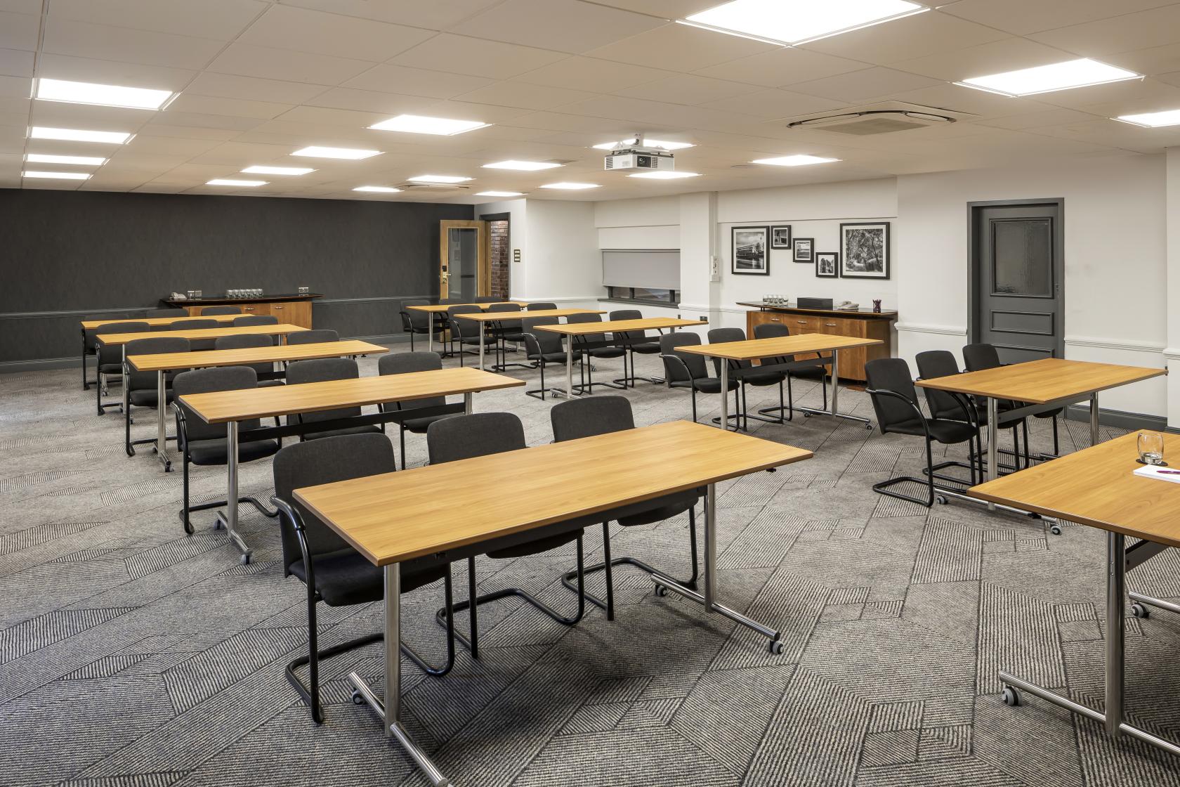 A meeting room with rectangular wood tables, black chairs, neutral carpeting, and framed photos at Crowne Plaza Nottingham Talbot Suite.