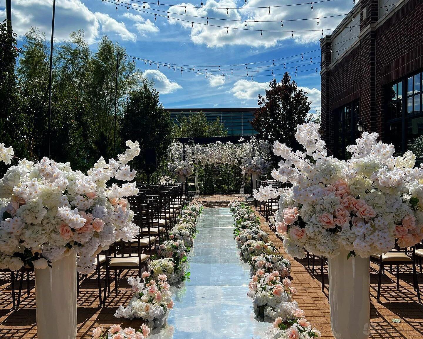 A terrace with a mirrored aisle, floral arrangements, and rows of chairs under string lights at Milano Event Center.
