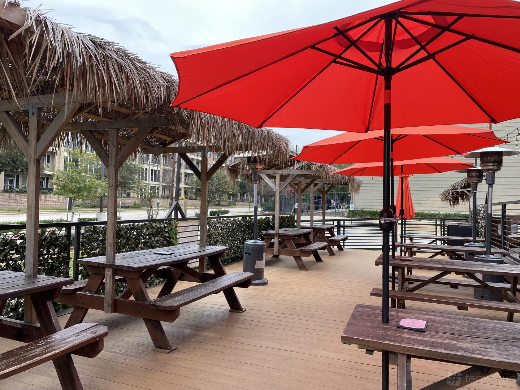 An outdoor restaurant patio with wooden picnic tables, red umbrellas, and palapa-style shade at Be More Pacific.