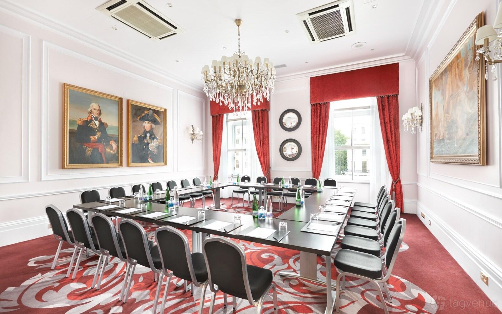 A meeting room with a U-shaped conference table, crystal chandelier, red curtains, and historic portraits at Clermont Charing Cross.