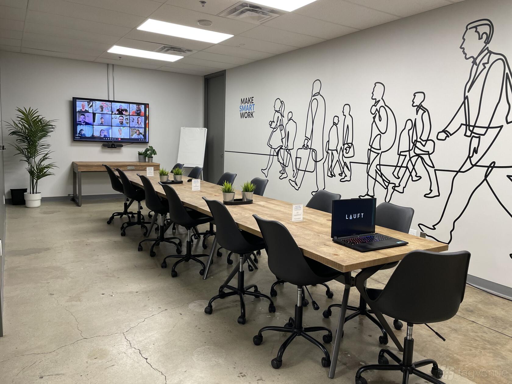 A boardroom with two long tables, black chairs, wall art of walking figures, and a video screen at LAUFT Burlington Centre.
