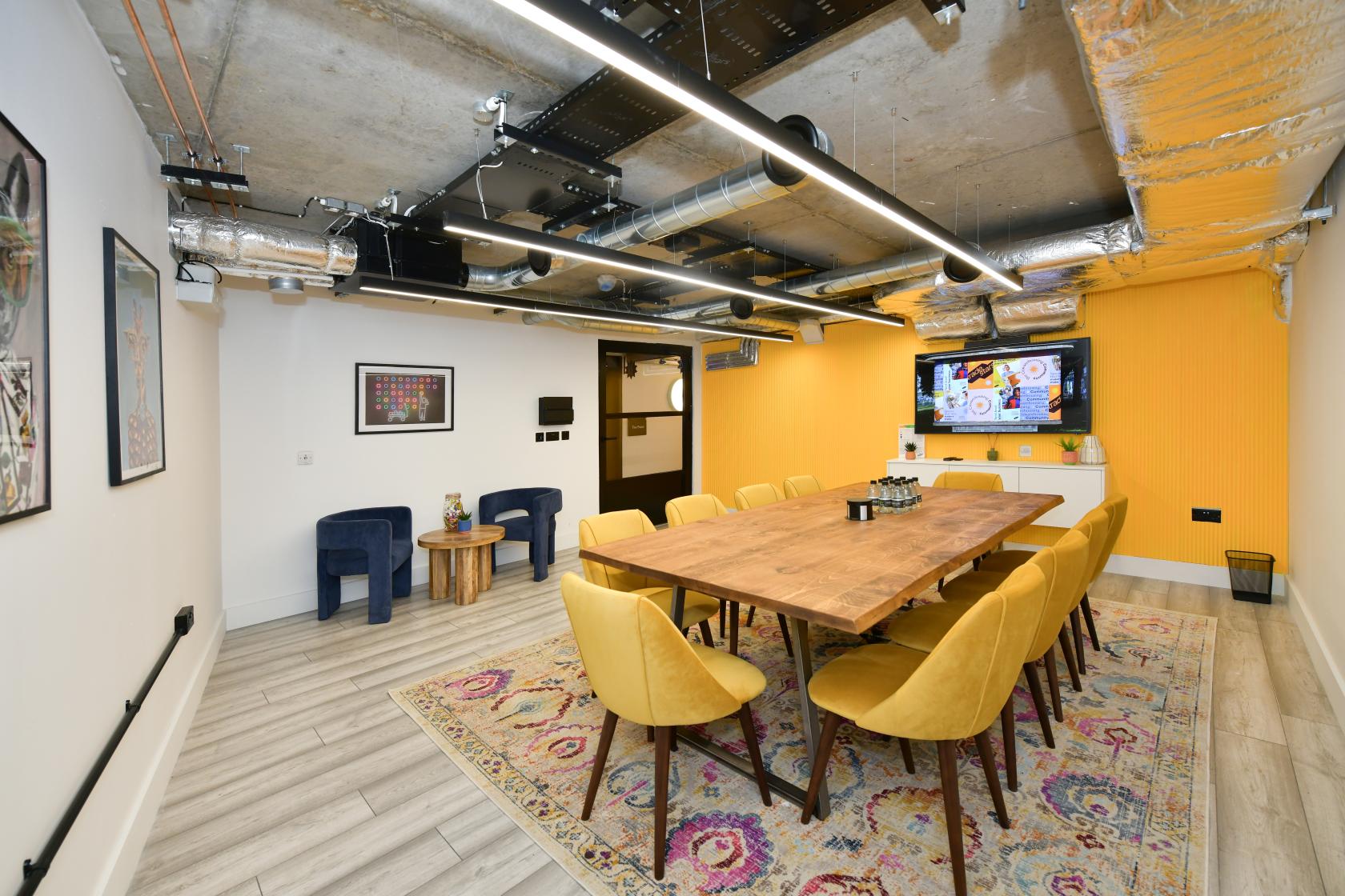 A meeting room with a long wooden table, yellow chairs, exposed ductwork ceiling, and wall-mounted screen at Tradestars Hackney Wick - Bookable Spaces.