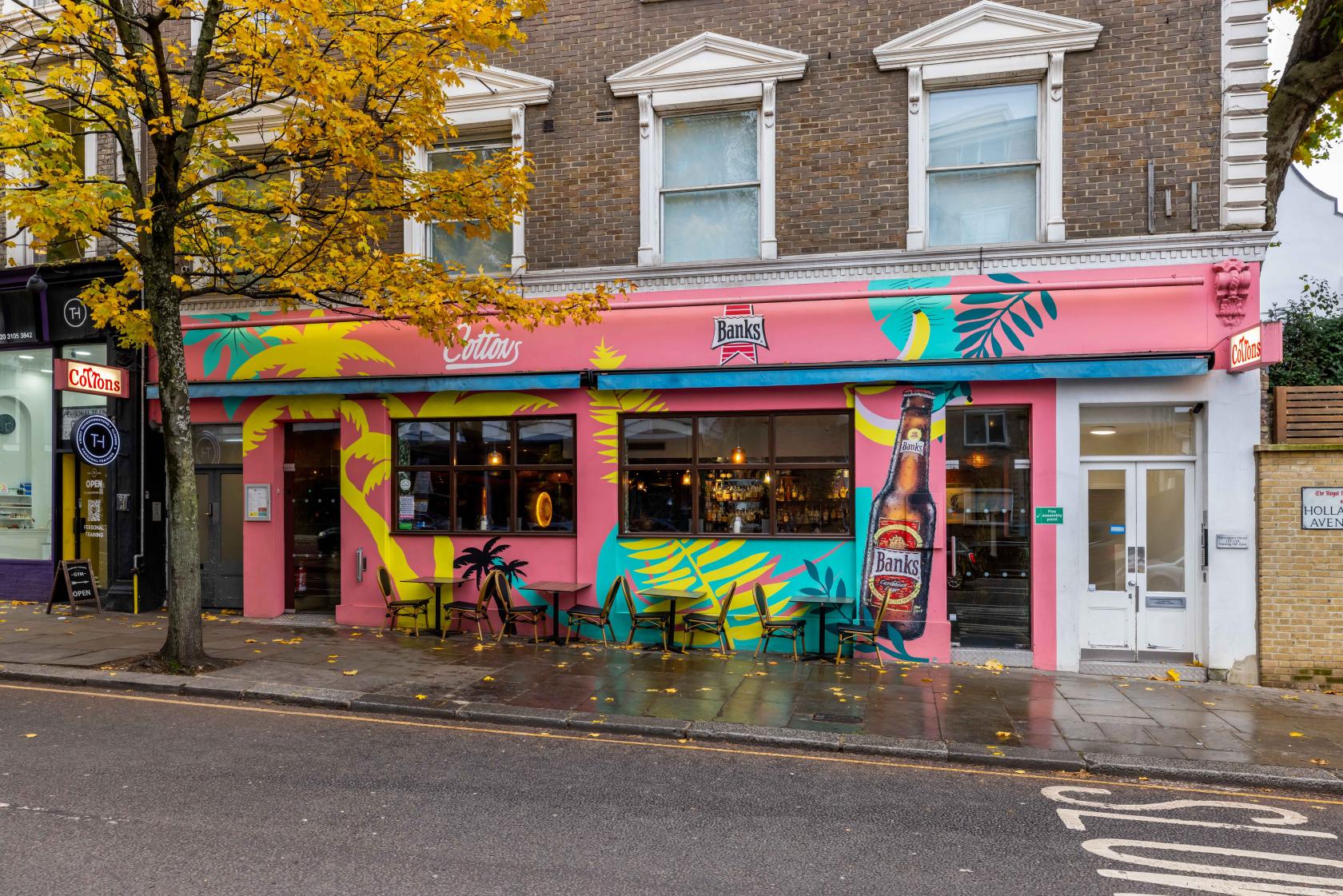 A restaurant with a vibrant pink and yellow mural, large street-facing windows, and outdoor seating at Cottons Notting Hill.