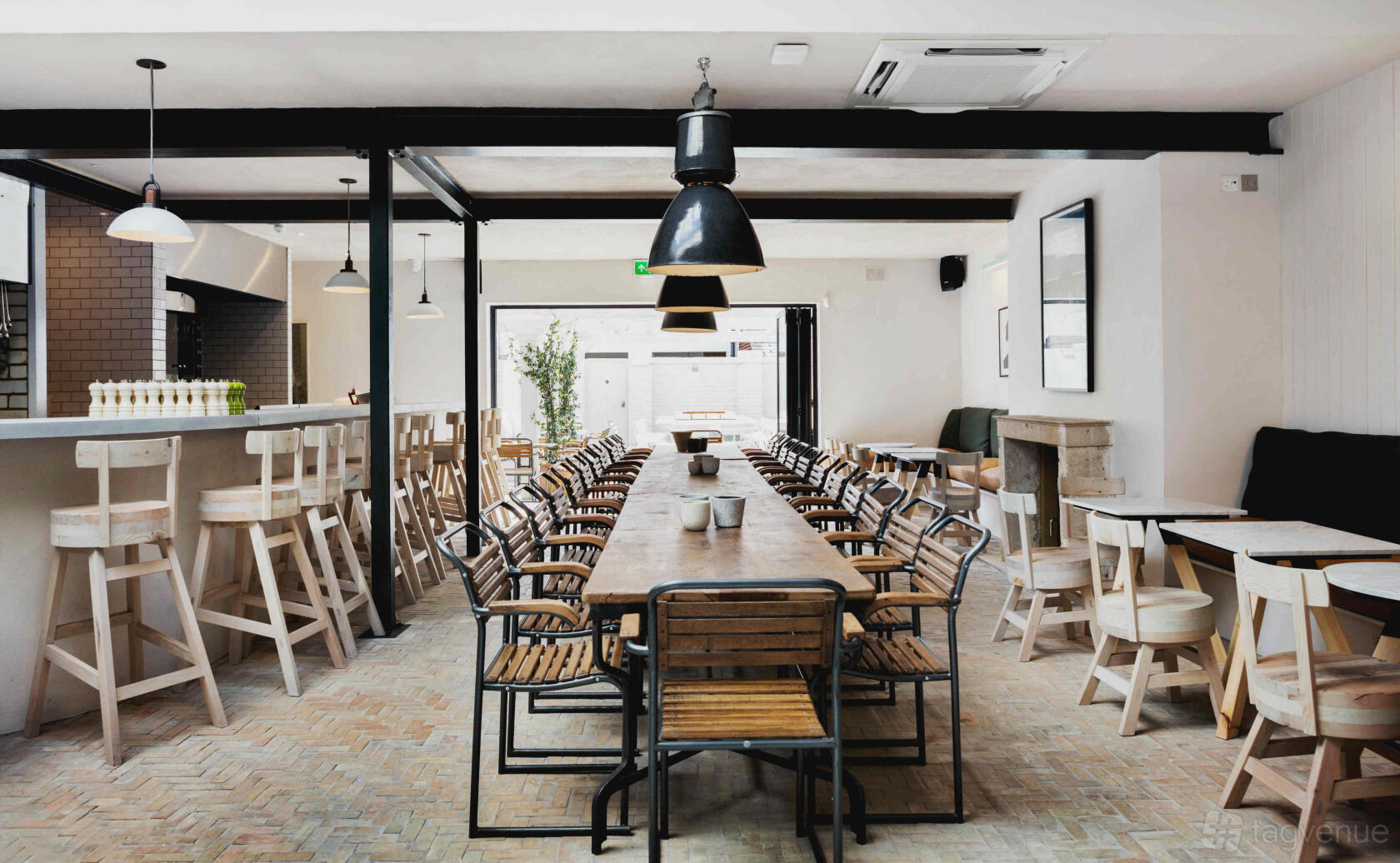 A bar with a long wooden communal table, metal chairs, pendant lighting, and brick flooring at No.197 Chiswick Fire Station.