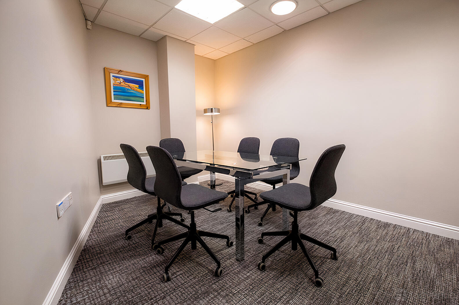 A meeting room with a glass table, six black swivel chairs, carpeted floor, and a framed wall art at Sky Business Centre Clontarf.