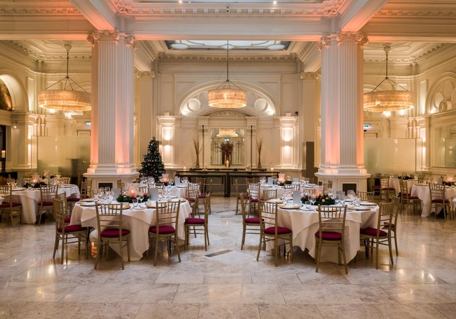A hotel ballroom with marble floors, tall white columns, chandeliers, and round tables set for dining at Andaz London Hotel.
