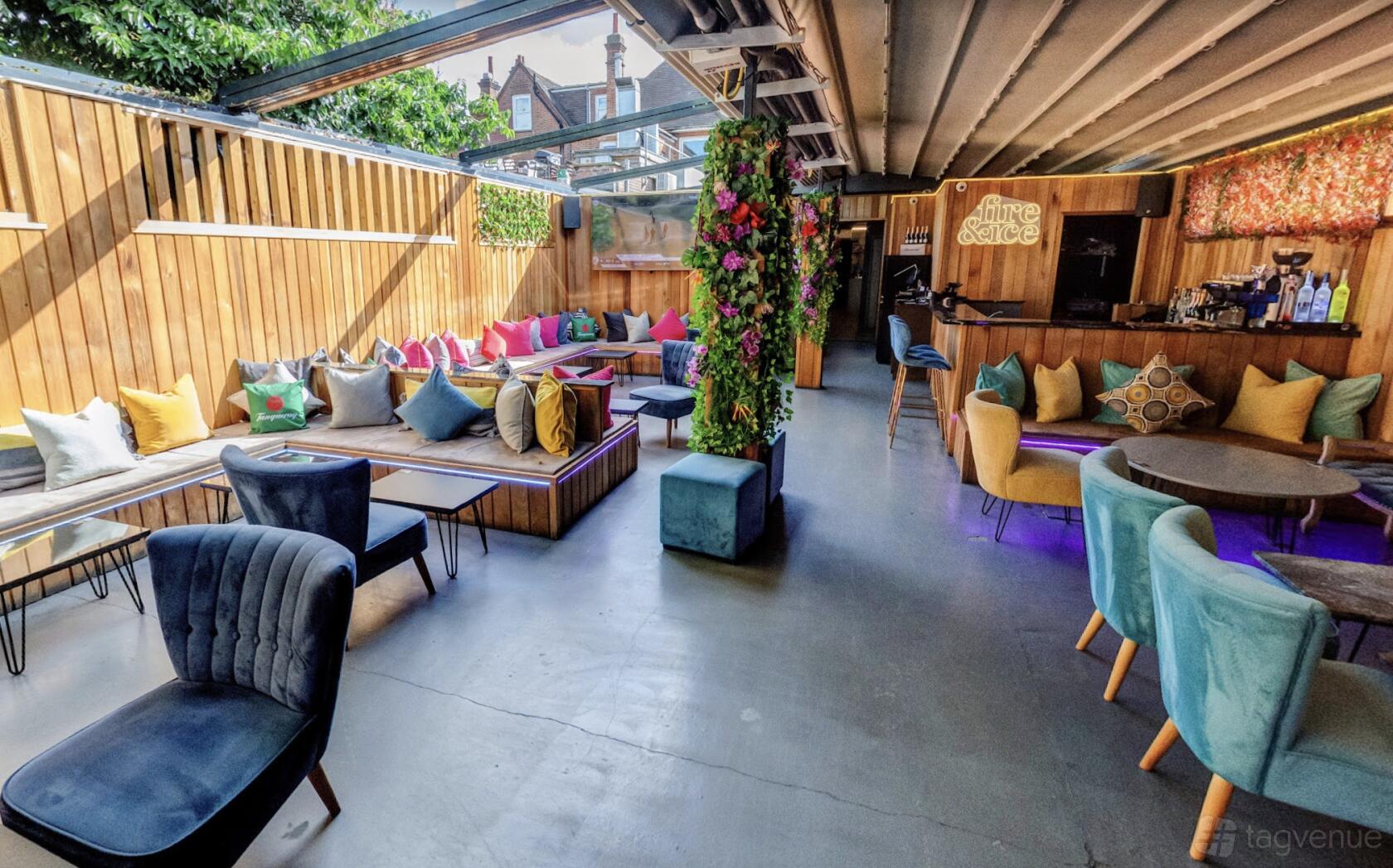 An outdoor lounge with colorful cushions, wooden seating, open roof panels, and greenery at Fire & Ice Lounge Bar.