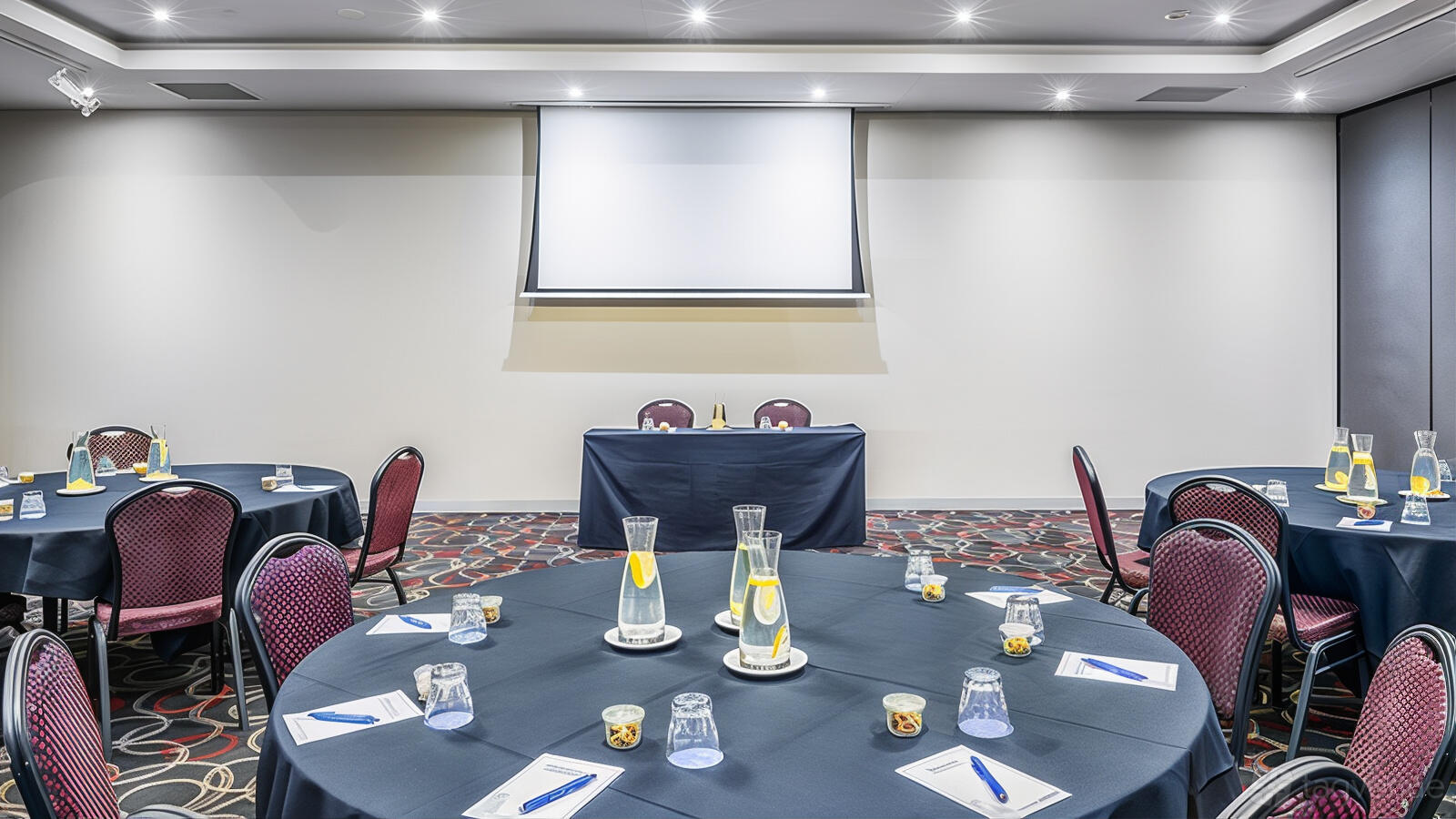 An event space with round tables, notepads, water pitchers, and a projection screen at Mantra Melbourne Airport.