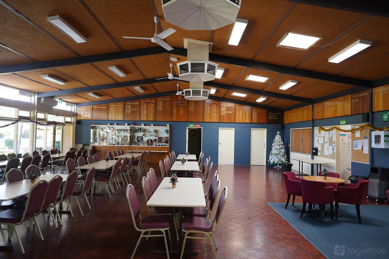 A spacious hall with long banquet tables, padded chairs, trophy cabinets, and a decorated Christmas tree at Blackburn Bowls Club.