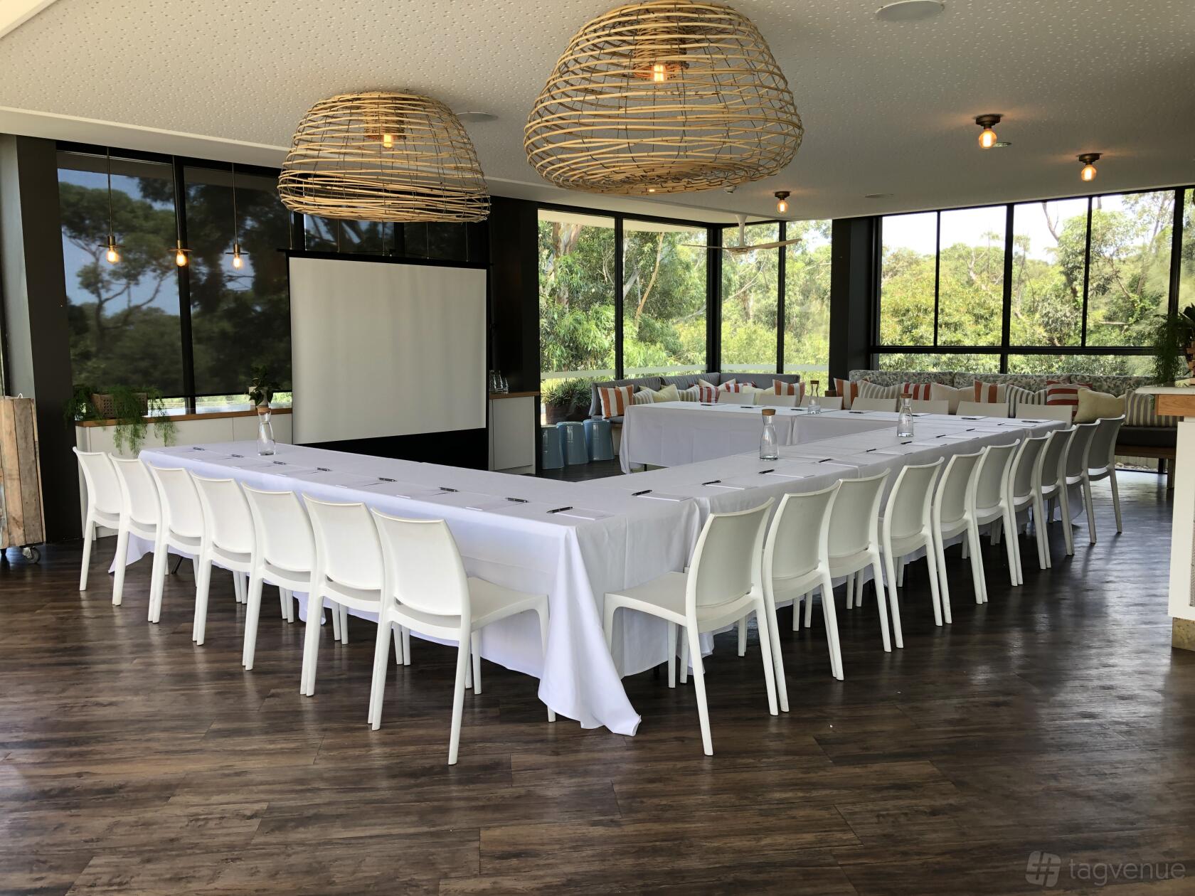 A rooftop event space with large windows, white chairs, U-shaped tables with linens, and rattan pendant lights at Bannisters Pavillion.