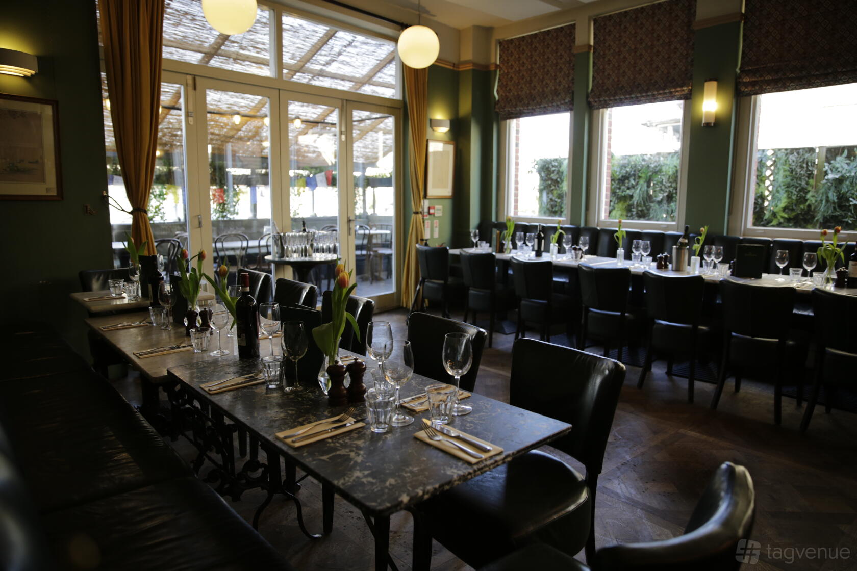A dining room in a pub with marble-top tables, black chairs, large windows, and set glassware at The Chamberlayne.