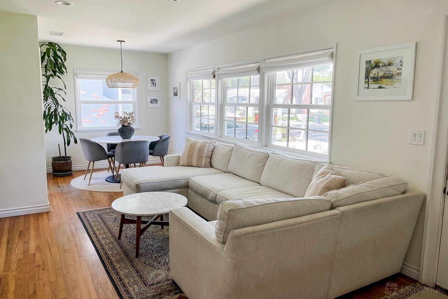 A house living room with a cream sectional sofa, round marble coffee table, and dining area by large windows at Bright and Airy Home.