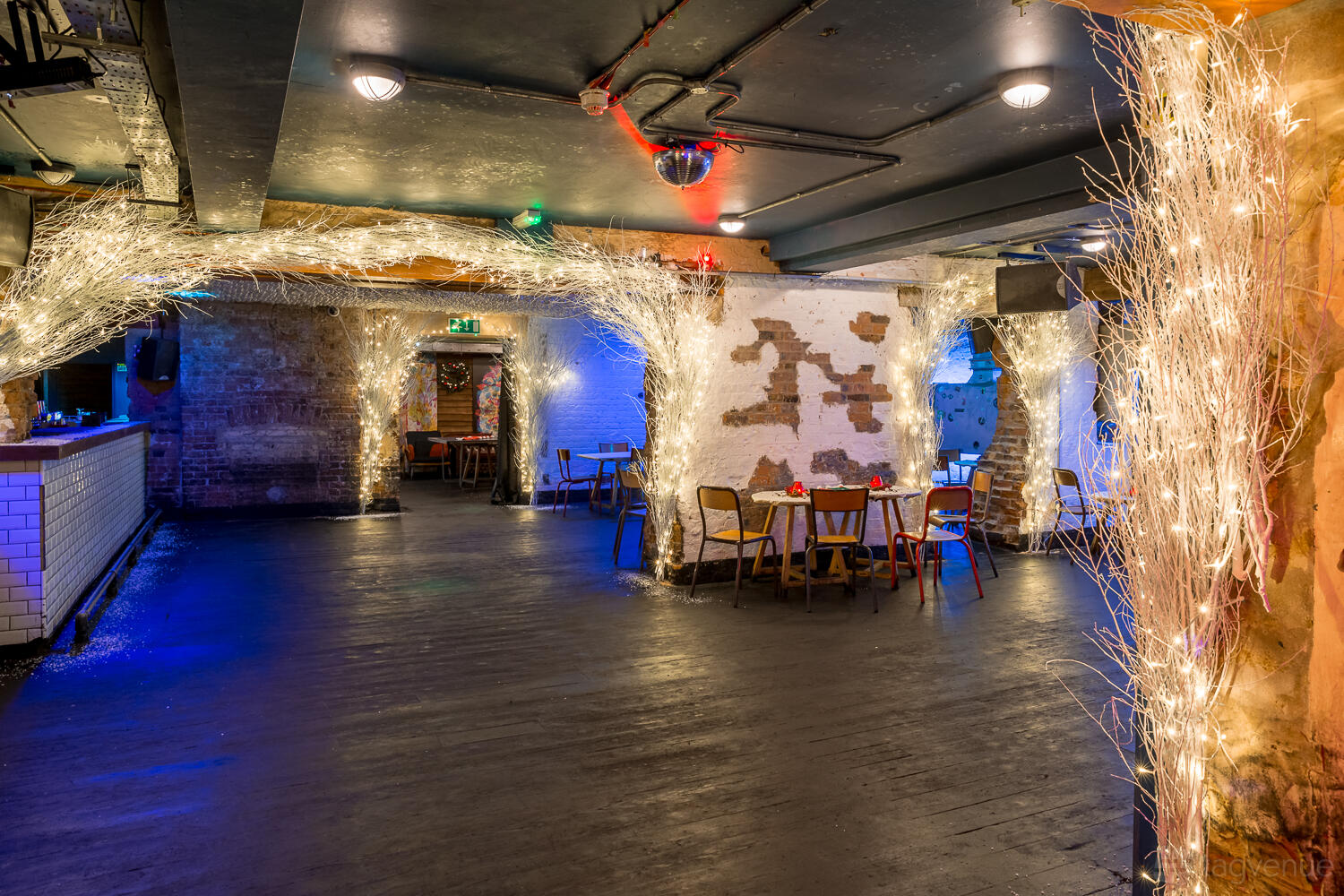 A basement bar with exposed brick walls, twinkling string lights wrapped around columns, and scattered tables at The Book Club.