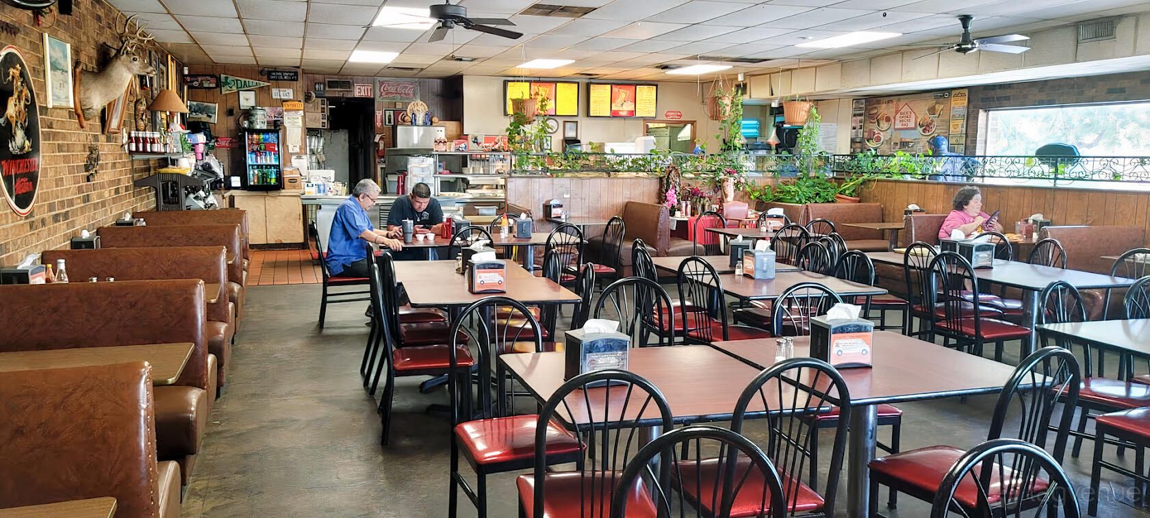 A restaurant dining area with wooden tables, red and black chairs, potted plants, and a counter at Rick's Smokehouse Barbecue.