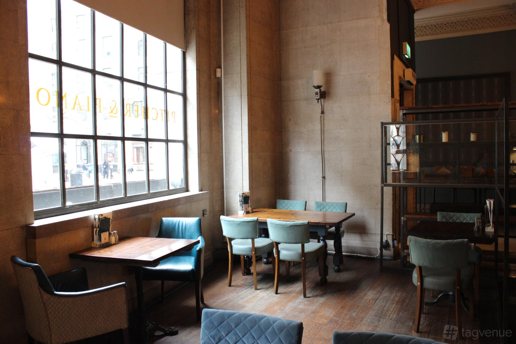 An event space in a bar with large windows, upholstered chairs, and wood tables at Pitcher & Piano Cornhill.